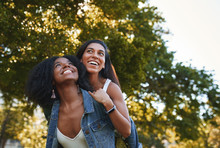 Portrait Of A Smiling Young African American Woman Giving A Piggyback Ride To Her Female Best Friend In The Park On A Sunny Day - Group Of College Students Laughing And Having Fun On Campus