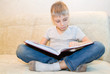 © sommersby - Little boy reads book sitting at home on beige sofa and is very interested in its contents