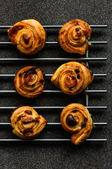 Wall Mural - Puff pastry snails with custard and raisins cooling down on a steel grate, deep grey background, shot from above