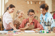 © AnnaStills - Young teacher in eyeglasses sitting at the table and showing how to draw a picture to her students at art studio