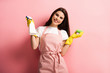 happy housewife in apron and rubber gloves holding spray bottle and sponge on pink background