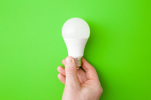 Young Man Hand Holding Led Light Bulb On Bright Green Table Background. Closeup. Energy Saving. Point Of View Shoot. Top Down View.