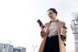 Closeup young Business Woman holding smartphone, Coffee to go and smiling. Attractive stylish Woman using her Phone near modern Building. Having Break. Smartphones. Games. People. Emotions. Business.