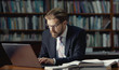 © MYDAYcontent - Busy businessman in formal suit and glasses typing on laptop keyboard sitting among books in library