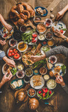 Fototapeta Uliczki - Turkish breakfast. Flat-lay of peoples hands taking and eating Turkish pastries, vegetables, greens, cheeses, fried eggs, jams and tea in copper pot and tulip glasses over wooden background, top view
