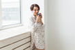 © satura_ - Calm and coziness. Beautiful young woman with cup of tea standing near the window at home