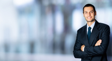 Wall Mural - Portrait of happy confident businessman in black suit and blue tie, with crossed arms pose, copy space for some text, advertising or slogan, standing against blured office background