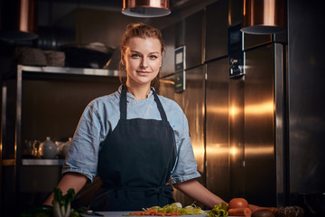 Confident and serious female chef standing in a dark kitchen next to cutting board with vegetables on it, wearing apron and denim shirt, posing for the camera, reality show look