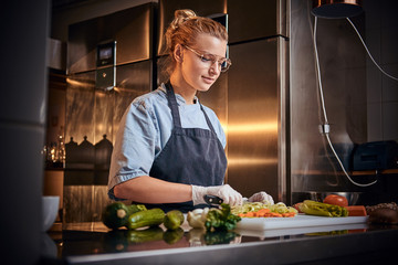 Wall Mural - Calm and focused female chef standing in a dark kitchen next to cutting board while cutting vegetables on it, wearing apron and denim shirt, posing for the camera, cooking show look