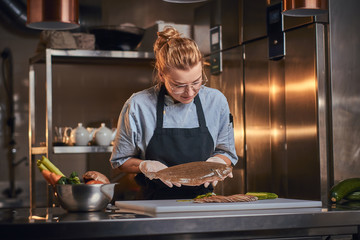 Wall Mural - Beautiful and stylish woman chef standing in a dark restaurant kitchen next to cutting board with vegetables on it, holding a big fish and wearing apron and denim shirt, posing for the camera, cooking