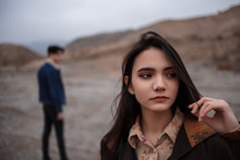 Dramatic Portrait Of A Young Brunette Girl In Cloudy Weather. Somewhere Behind Her, Out Of Focus, Her Young Lover Boyfriend Leaves Her After Break Up . Selective Focus, Small Focus Area