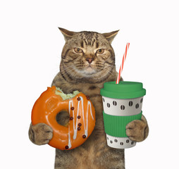 The beige cat is holding an orange bitten donut and a blue paper cup of coffee with a plastic lid and a straw. White background. Isolated.