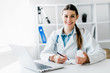 © zadorozhna - Smiling young female doctor working at the clinic , she is using a computer and writing medical reports.