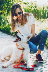 Wall Mural - Smiling caucasian girl dressed in blue jeans and white t-shirt is playing joyfully with her dog in a the light of the sun