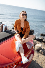 Wall Mural - Portrait of a stylish woman sitting on the car hood, while traveling by car near the ocean. Concept of a summer time and carefree travel