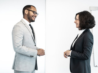 Two friendly confident professionals discussing business issues in conference room. Business man and woman standing indoors near white board and chatting. Business communication concept