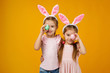 © producer - happy cute little child girls with pink bunny ears holding painted Easter eggs on studio yellow background. Easter day