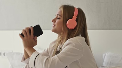 Wall Mural - An attractive young happy woman in pajama is listening to the music with cellphone and headphones while singing on bed in light room