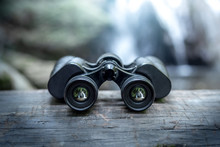 Close Up Binoculars On Wood In Forest With The Waterfall Behind.