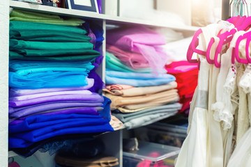 Rack with many beautiful holiday dresses for girls on hangers and multicolored fabric stack at children fashion sewing studio and showroom indoor. Female dress rental salon maker photo session event