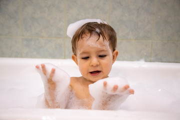 Wall Mural - Cute baby boy taking a bath looking at soap foam in his hands. close up, soft focus, bacground is bathroom in blur