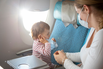 Cute little toddler girl putting on a medical respiratory mask in an airplane chair. sitting next to a young mother in a mask. rest in asia. coronavirus epidemic. covid-19