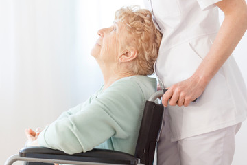 Bright photo of positive elderly woman on wheelchair with her helpful volunteer at nursing facility