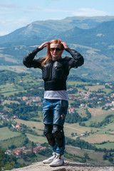 Girl dressed in a motorcycle outfit. body protection turtle and knee pads on the edge of the top of the mountain. incredible view. Motorcycle travel concept. Vertical photo. San Marino