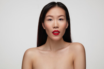 Charming brown-eyed pretty young brunette lady with red lips showing her white perfect teeth and looking calmly at camera, standing over white background