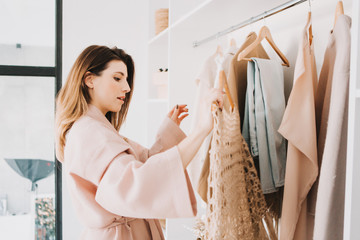 Cute young woman standing in front of hanger rack and trying to choose outfit.