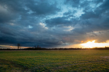 The Glow Of The Sun Coming Out From Behind A Dark Cloud Over A Green Meadow, Evening View