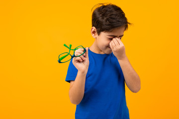 Wall Mural - dry eye syndrome. a boy in a blue T-shirt holds glasses in his hand and rubs his eyes on a yellow background with copy space