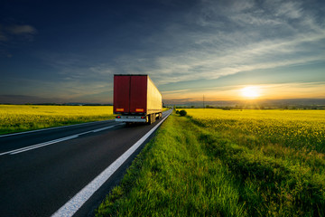 Wall Mural - red truck departing into the distance on the asphalt road between the yellow flowering rapeseed fiel