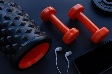 Two orange dumbbells, weight plates, white in ear headphones and bumpy foam roller on gym floor