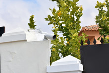 Surveillance Camera On The White Exterior Wall Of A House And Next To A Fence With Spikes