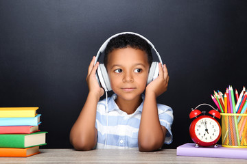 Wall Mural - Young African American school boy sitting at desk with books, pencils and headphones on black background