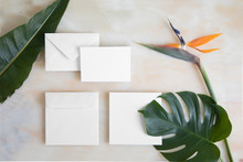 Empty Card With Envelope On Marble Table And Tropical Flowers. Mockup Template With Top View. Strelitzia Flowers And Monstera Leaves On Marble Floor.