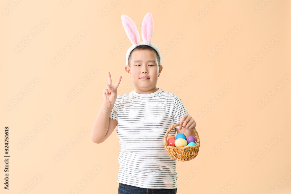 Little Asian boy with Easter eggs and bunny ears showing victory gesture on color background