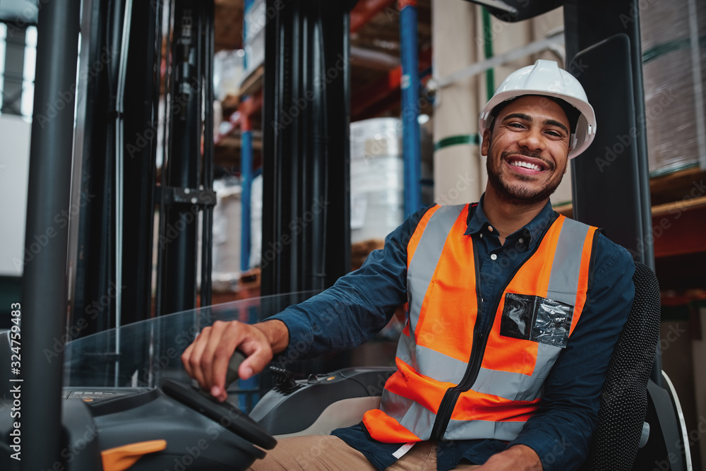 Young forklift driver sitting in vehicle in warehouse smiling looking at camera - obrazy, fototapety, plakaty 