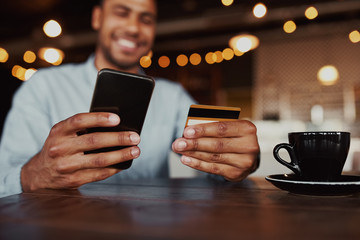 Closeup of cheerful happy african man hand holding mobile phone typing card data to make online payment sitting in cafeteria