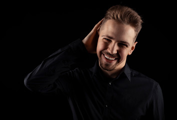 Cheery young man with goatee in black shirt touching hair with one hand looking at camera, isolated