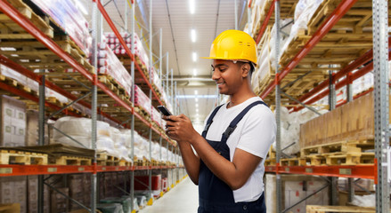 profession, logistic business and technology concept - happy smiling indian worker or loader in helmet using smartphone over warehouse background