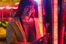 Happy Asian Girl Using Smartphone At Amusement Park - Young Trendy Woman Having Fun With Tecnology Trends With Neons In Backgrounds - Tech And Youth Concept - Soft Focus On Face