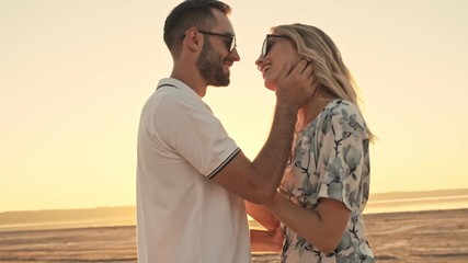 Poster - A stylish young couple man and woman wearing sunglasses are hugging on the sunny beach near the lake