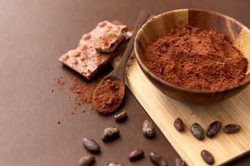 Wall Mural - sweets, confectionery and culinary concept - chocolate with hazelnuts, cocoa beans and powder in wooden bowl with spoon on brown background