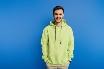 cheerful handsome man in hoodie smiling at camera while holding hands in pockets isolated on blue