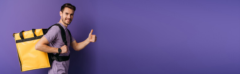 panoramic shot of happy delivery man showing thumb up while looking at camera on purple background