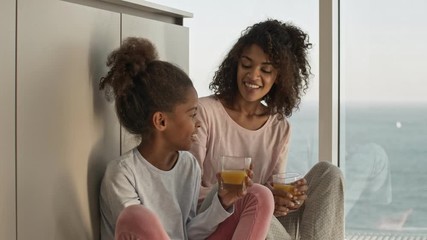 Wall Mural - Happy african woman and her pleased little pretty daughter drinking juice while sitting on the floor near window at home