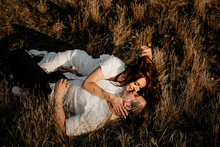 Loving Couple Laying And Kissing On The Grass At Sunset In Summer. Brunette Girl In A Light Dress. Love Story.
