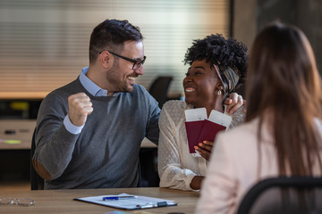 Wall Mural - Young businesscouple choosing a trip with agent sitting at the travel agency office. Happy clients with passport. Adventure concept. Young couple visiting travel agency